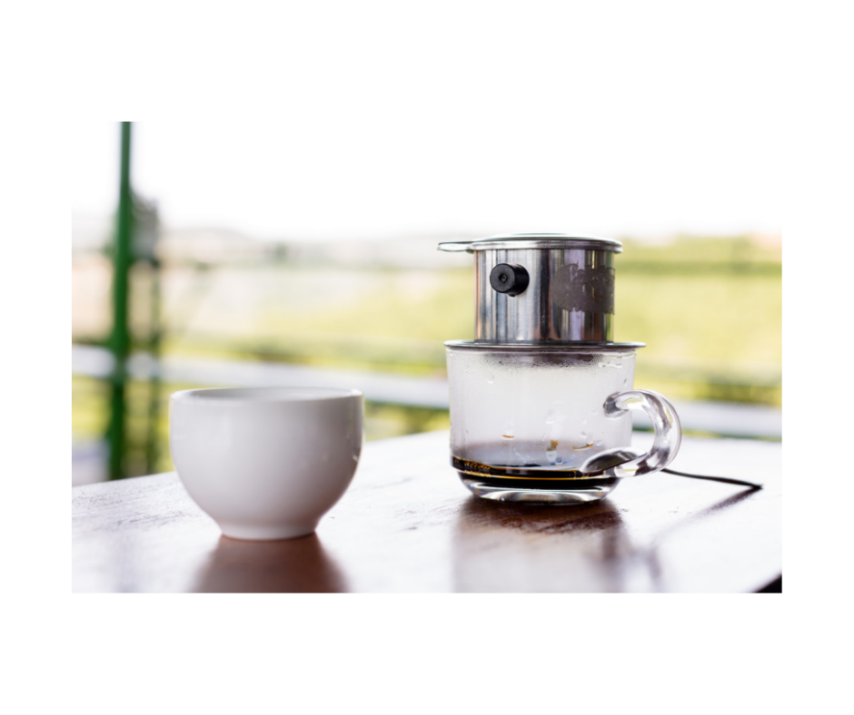 auf einem Serviertisch ein Glas, darauf ein vietnamesischer Kaffeefilter, ein "Phin". Der Kaffee tröpfelt durch. Daneben ein Schälchen mit Eiswürfeln